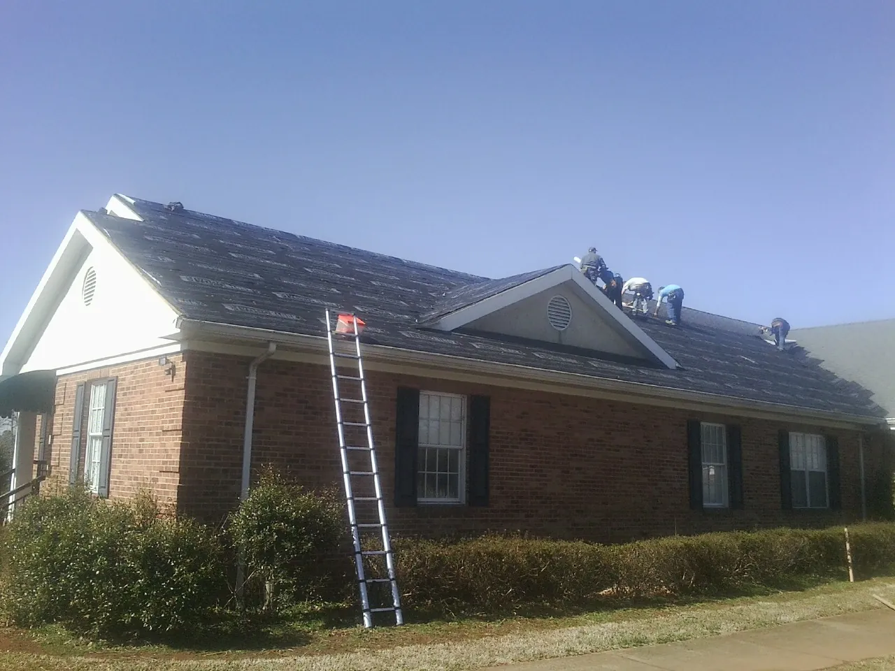 Crew tearing shingles off a brick ranch roof with a ladder set — Metal Roof Repair work by our crew in Admire