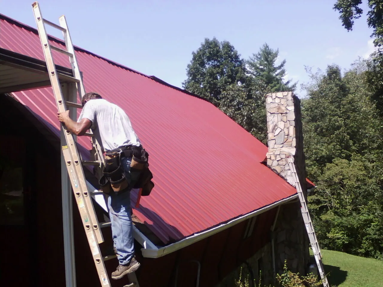 Roofer on a ladder beside a red metal roof and stone chimney — Soffit & Fascia Repair by Roofing Brothers in Admire